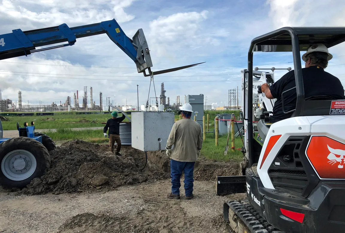 Technicians and a telehandler lowering a precast concrete vault into a trench at an industrial facility