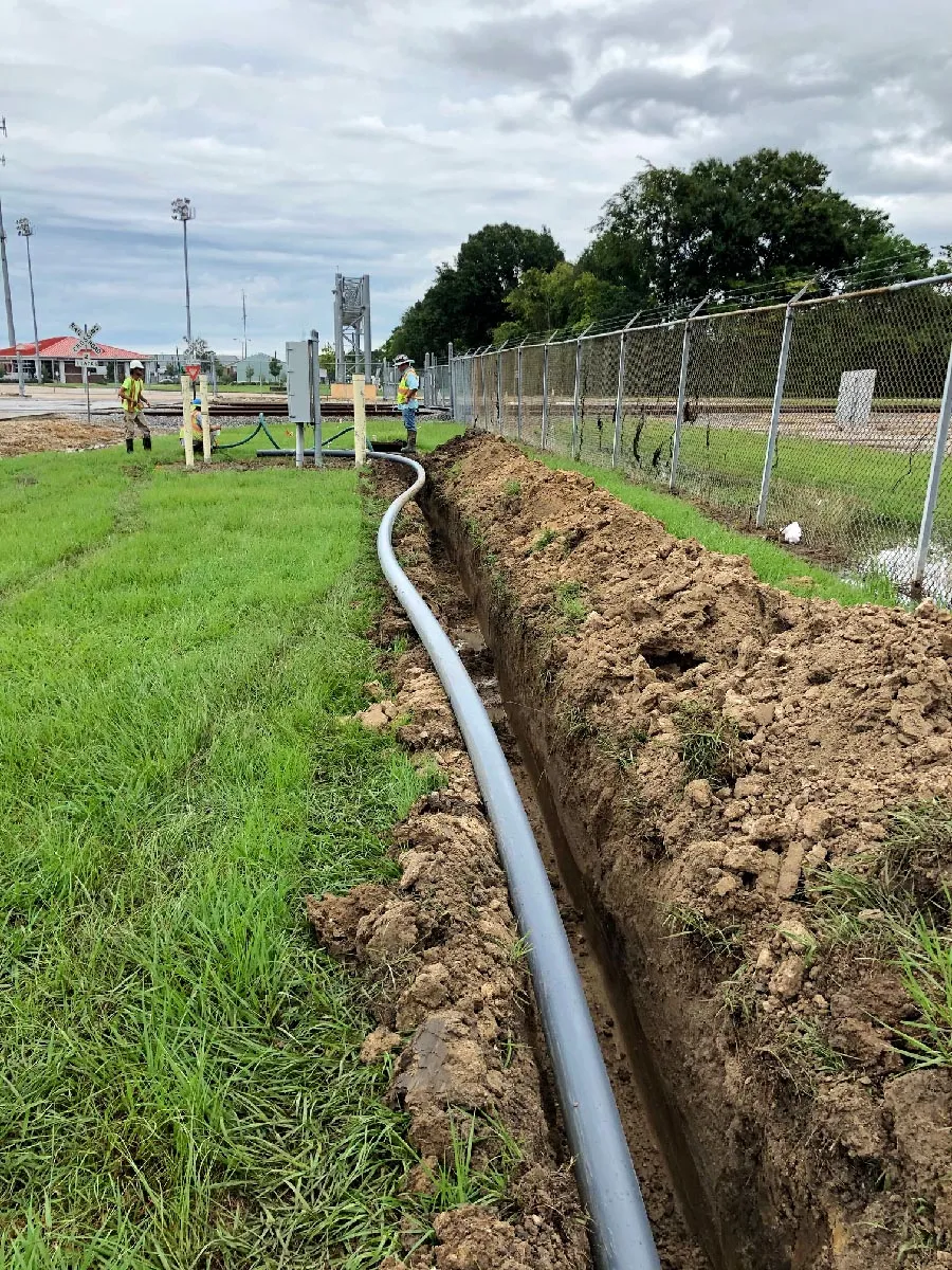 Technicians laying conduit in an open trench along a fence line near railroad tracks
