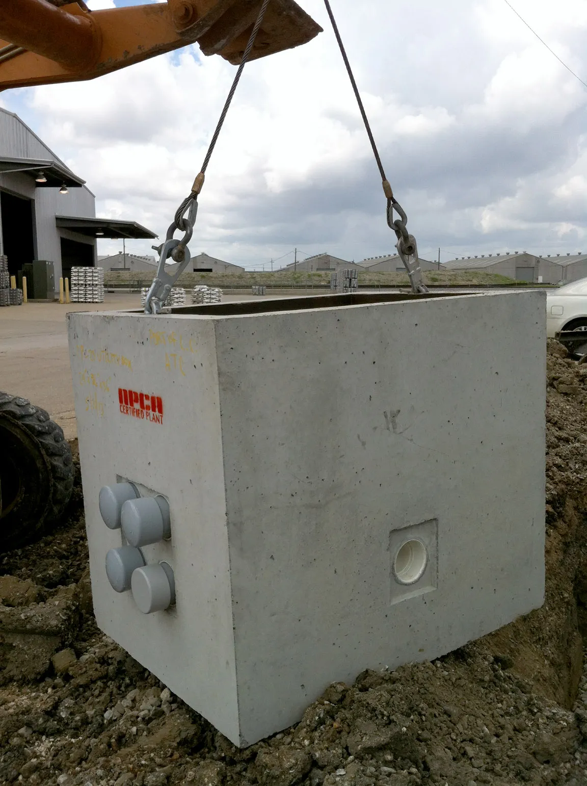 Precast concrete vault being lifted by an excavator's cable sling at an industrial site