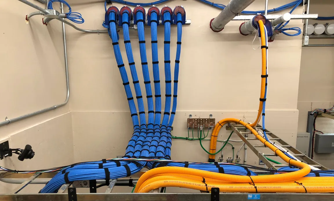 Blue cable bundles fanning from an overhead tray down to a wall-mounted patch panel and grounding bar